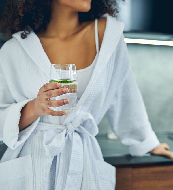 Een vrouw in een witte badjas drinkt een glas water in de keuken